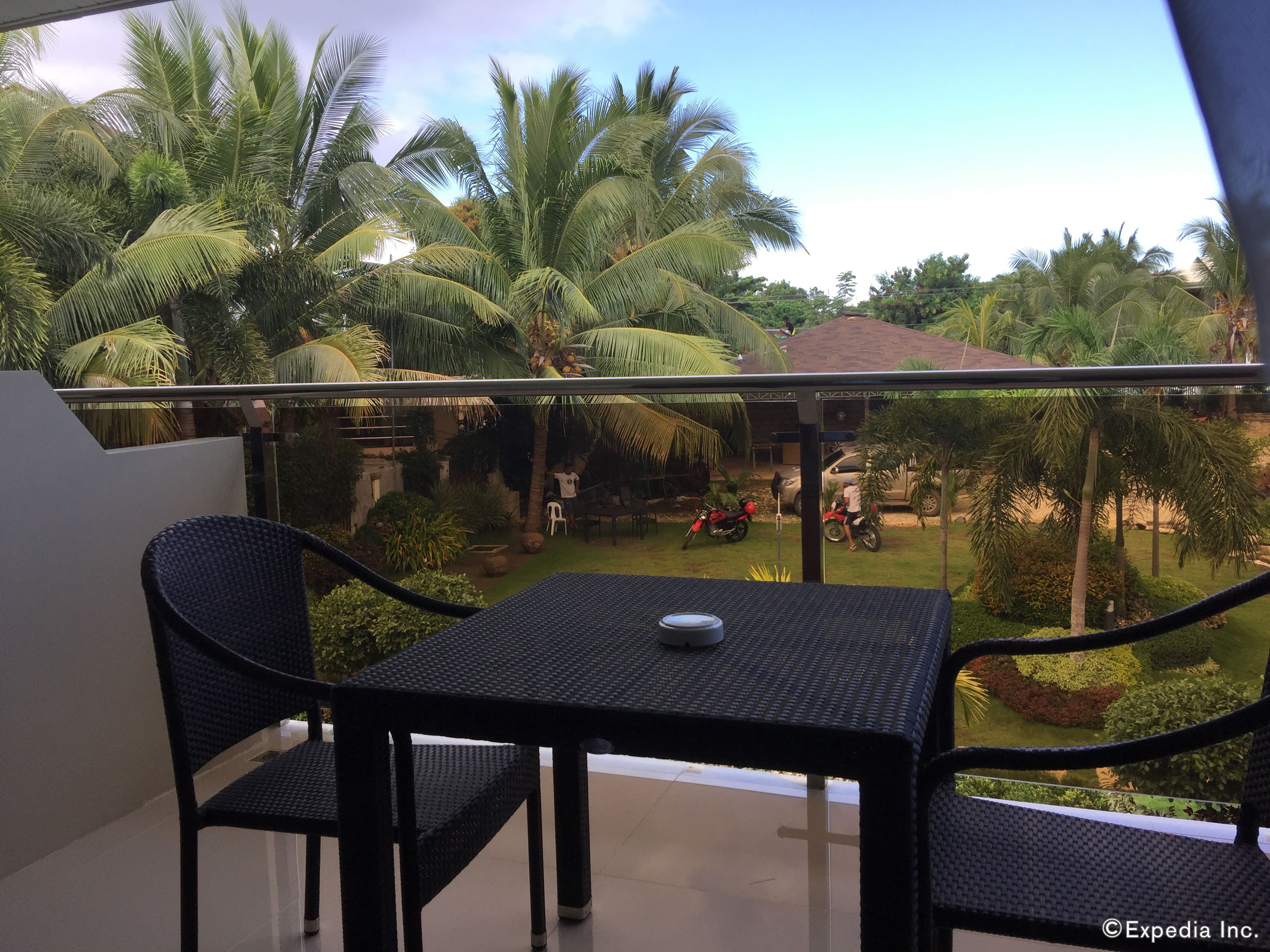 A balcony with a table and chairs, overlooking a garden with palm trees and a parked motorcycle.