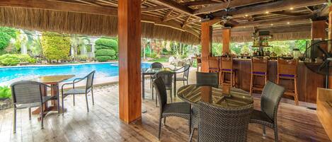 An outdoor bar area with wooden flooring, wicker furniture, and a pool in the background.