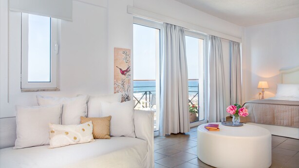 A modern hotel room with a white sofa, a round coffee table with books and flowers, a bed with a headboard, and large windows with curtains. - Aqua Marina (Rethymno)