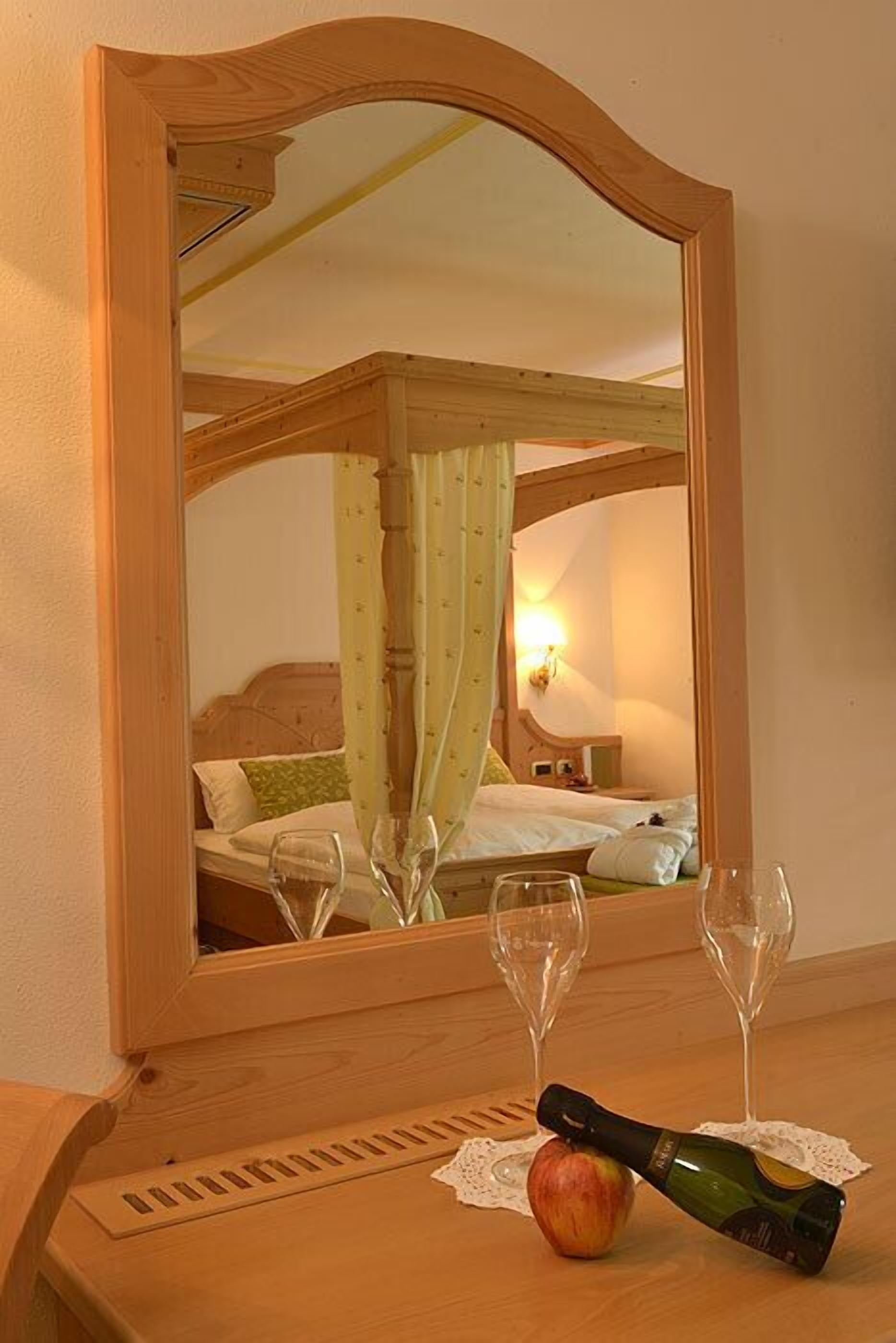 Een houten spiegel reflecteert een slaapkamer met een bed, een nachtkastje met twee wijnglazen, een fles en een appel.