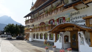 Un hotel de estilo arquitectónico alpino tradicional, con balcones adornados con jardineras y una señal que dice "Hotel Andreas Ristorante", con vistas a montañas al fondo.