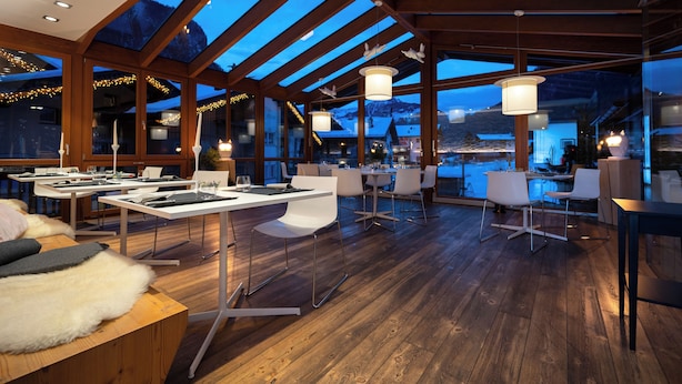 Amplio espacio interior con suelo de madera, mesas, sillas y ventanas grandes que ofrecen vistas a una montaña nevada.