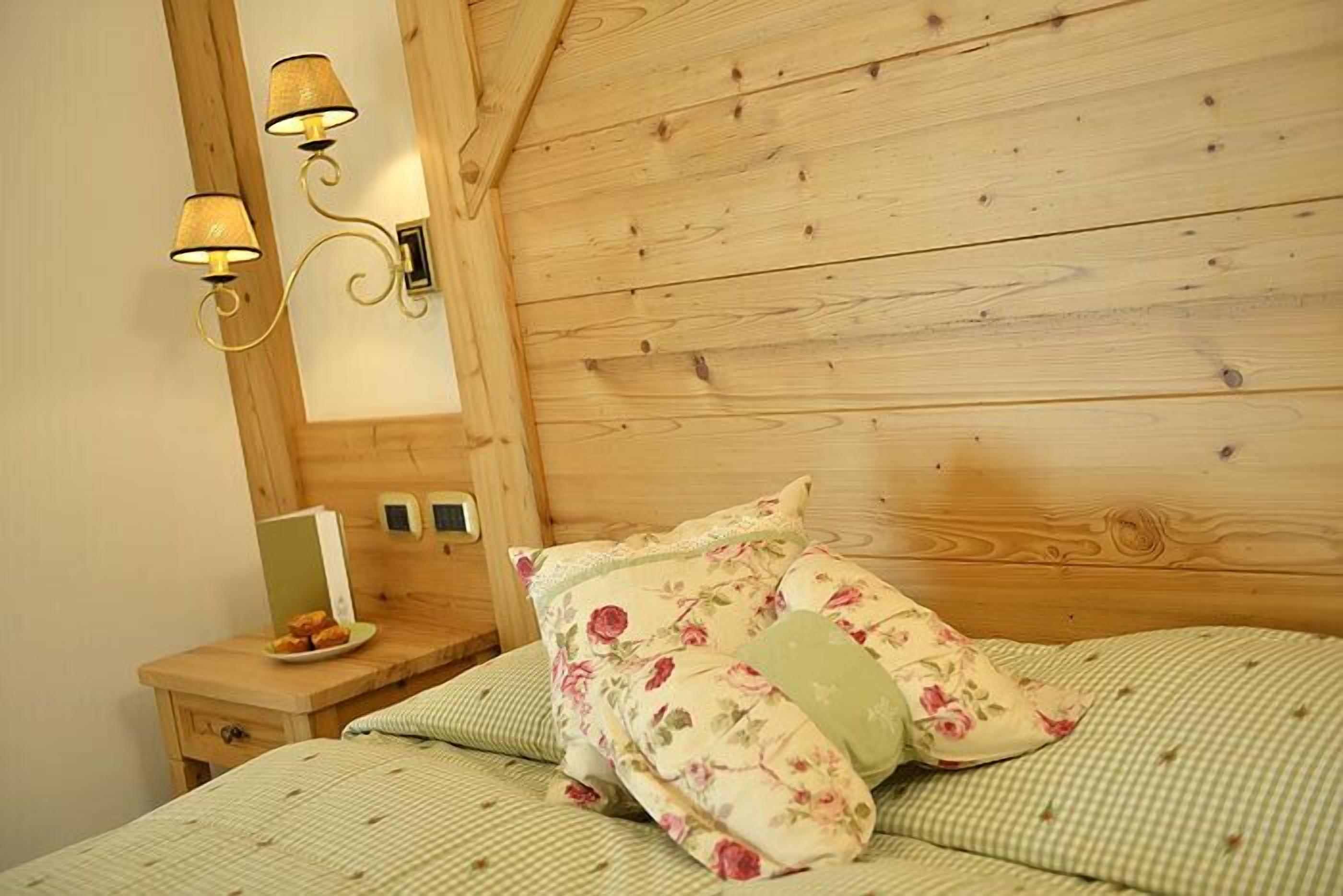 A wooden-paneled bedroom with a bed, floral pillows, a bedside table with a lamp, and a mirror.