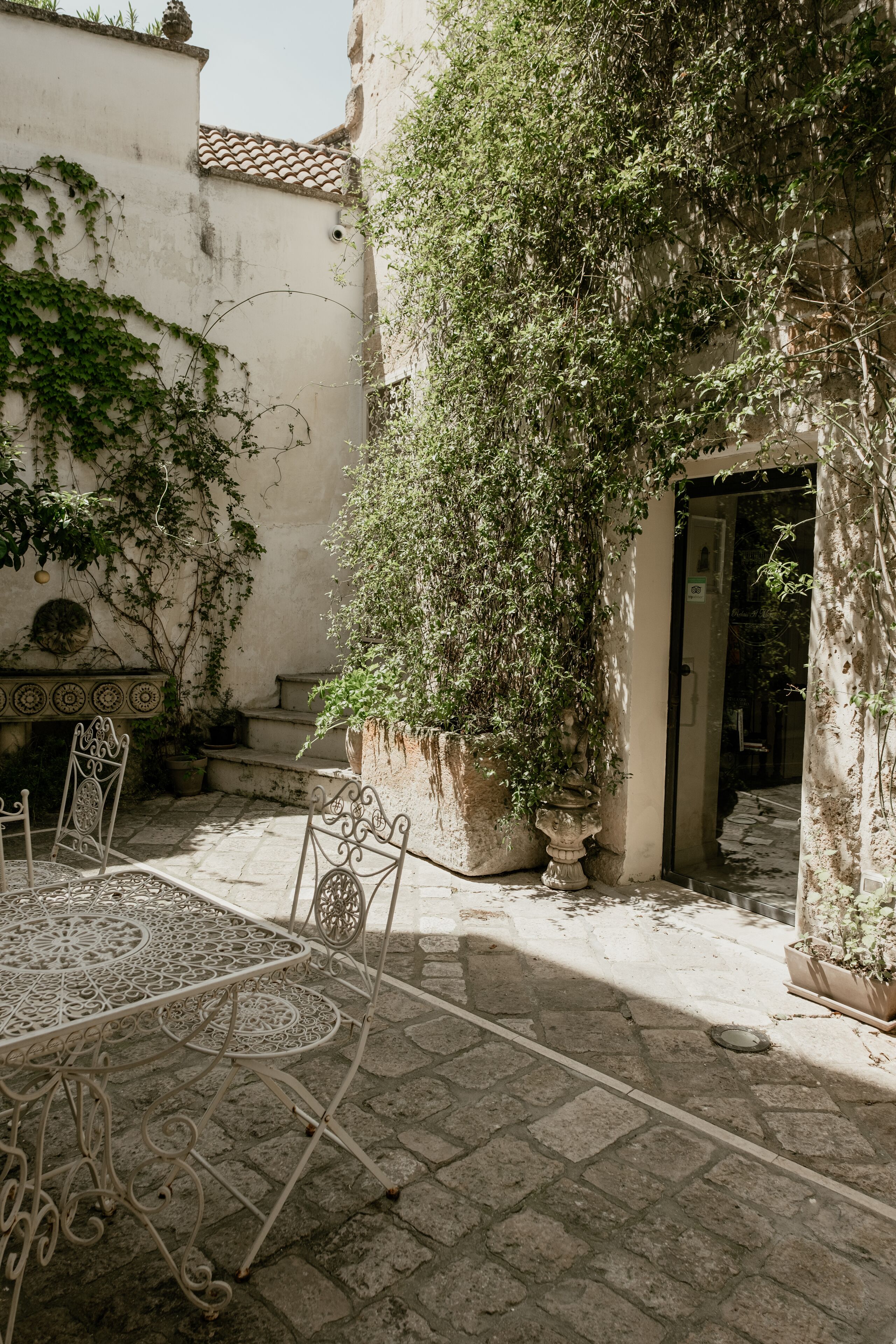 An outdoor patio with a wrought iron table and chairs, surrounded by potted plants and a stone wall.