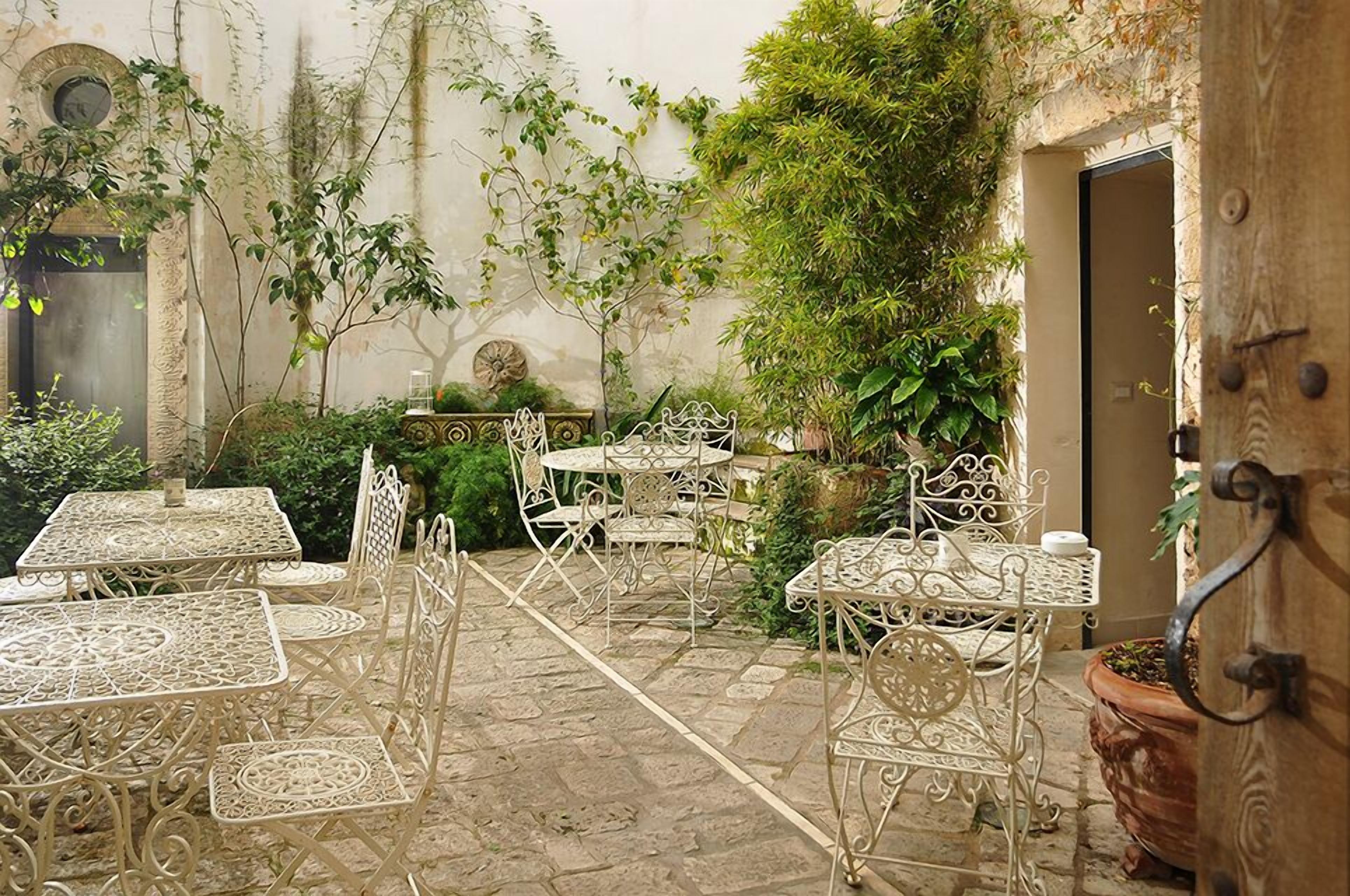 An outdoor patio with metal tables and chairs, surrounded by greenery and architectural details.