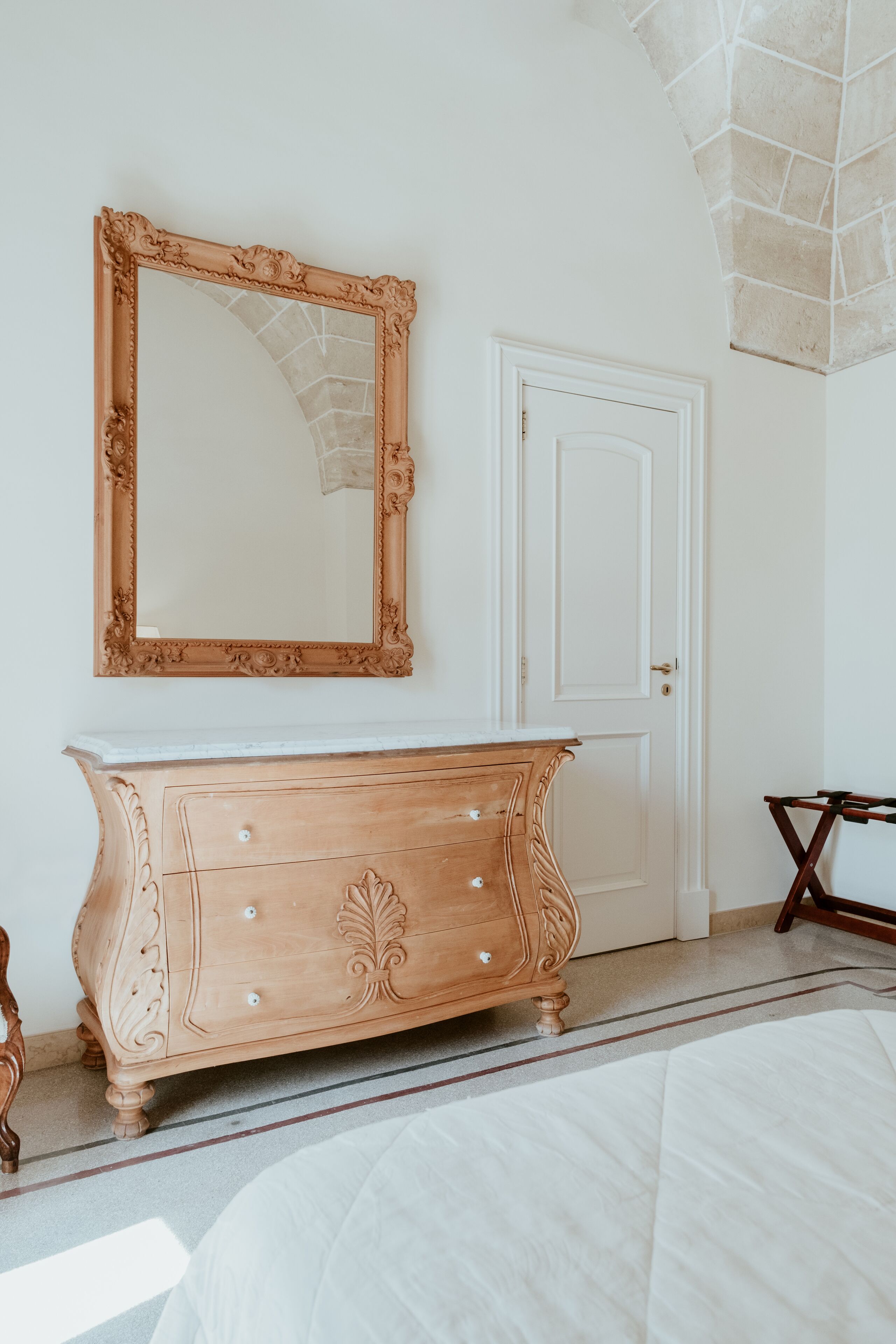 Una stanza con un comò in legno, uno specchio grande, una porta bianca e un letto con copripiumino bianco.