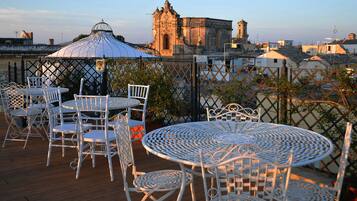 A terrace with white metal tables and chairs, overlooking a historic building and a river.
