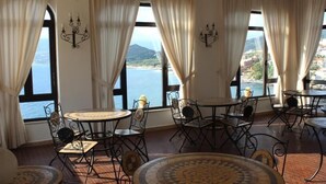 A dining area with tables and chairs, large windows with curtains, and a view of the sea and hills. - Hotel Palinuro (Centola)