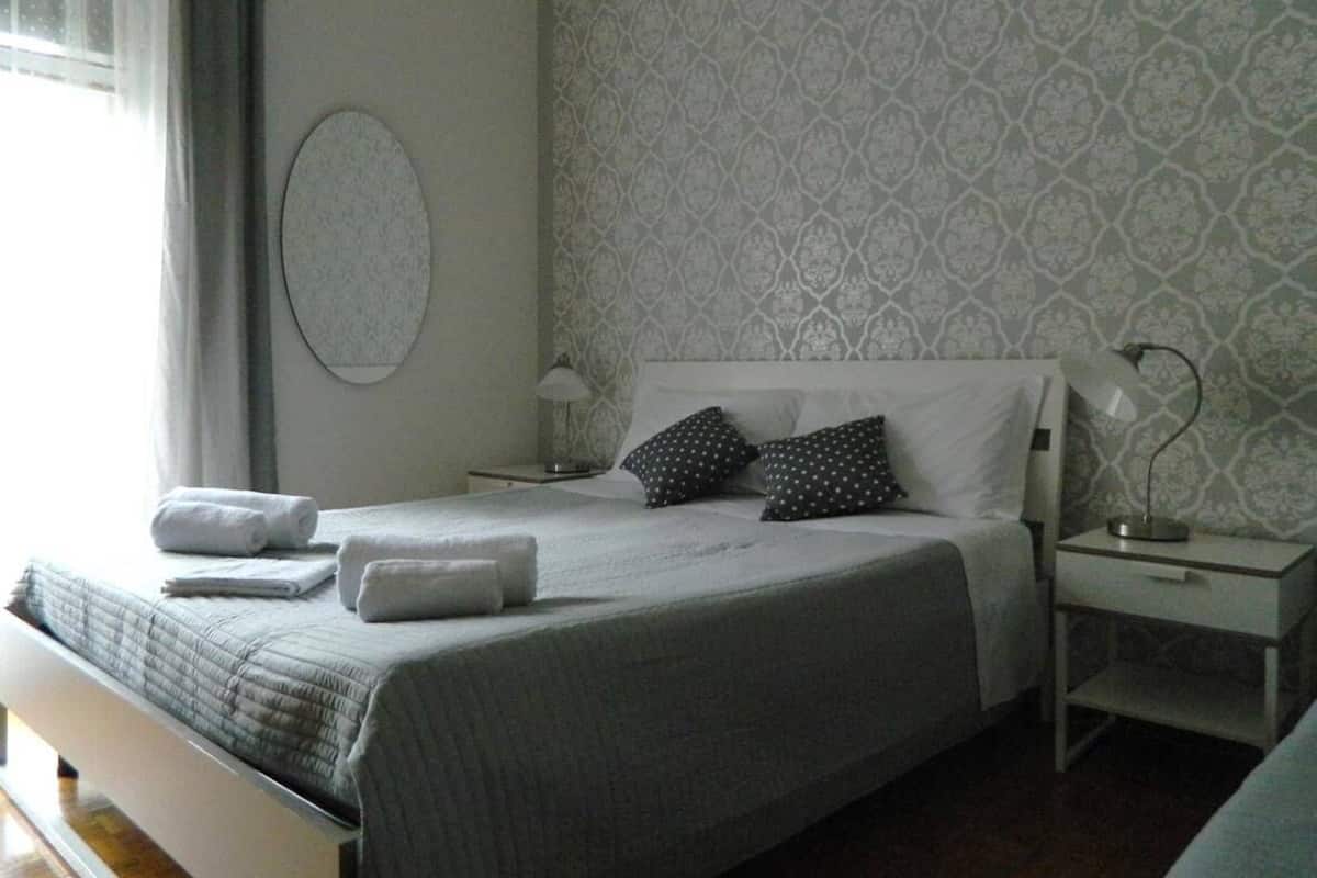Una cama bien hecha con almohadas y una toalla doblada, una mesita de noche con lámpara y un papel tapiz estampado en la pared.