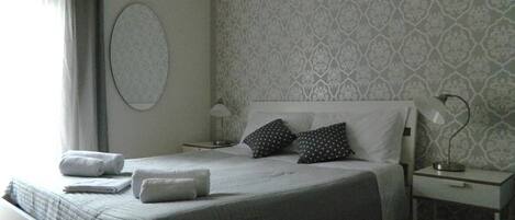 Una cama bien hecha con almohadas y una toalla doblada, una mesita de noche con lámpara y un papel tapiz estampado en la pared.