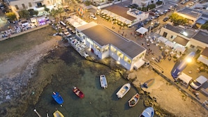 An aerial view of a coastal town with a prominent white building with large windows, a sandy beach with several small boats, and a bustling street with people and shops. - CiCò Hotel (Carovigno)