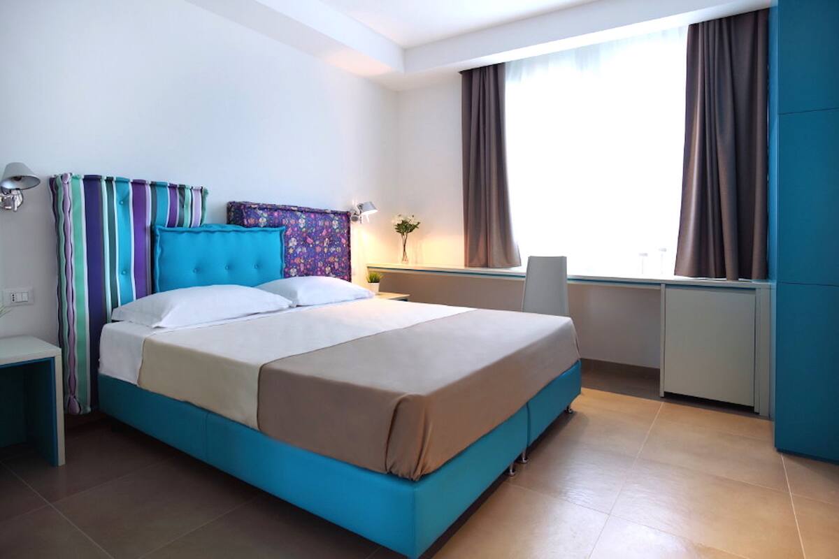 A hotel room with a bed, bedside table, lamp, and a desk with a chair.
