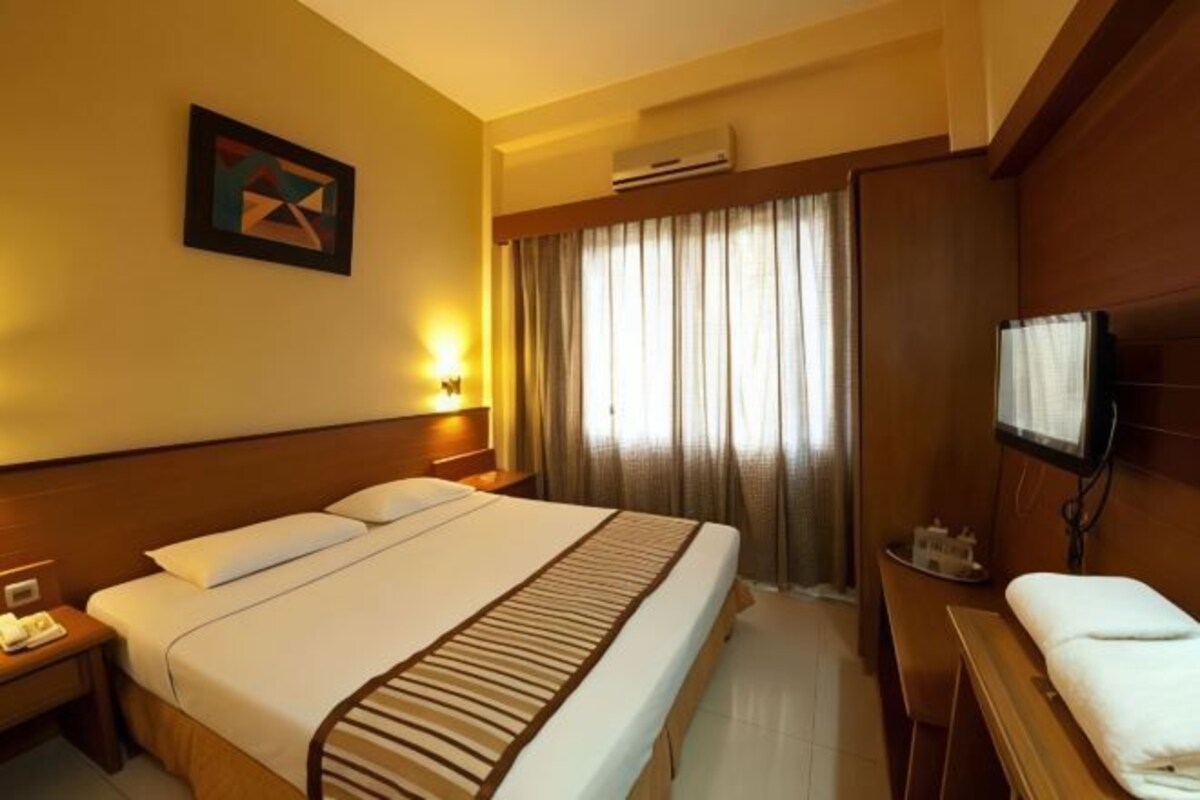 Ein Hotelzimmer mit einem großen Bett, einem Nachttisch mit Lampe, einem Fernseher und einem Fenster mit Vorhängen.