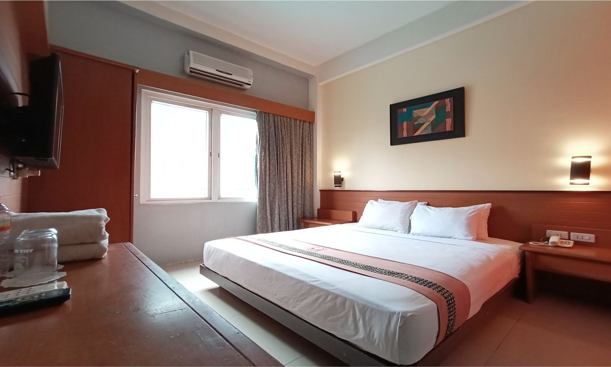 A hotel room with a large bed, a television mounted on the wall, an air conditioning unit, and a painting on the wall.