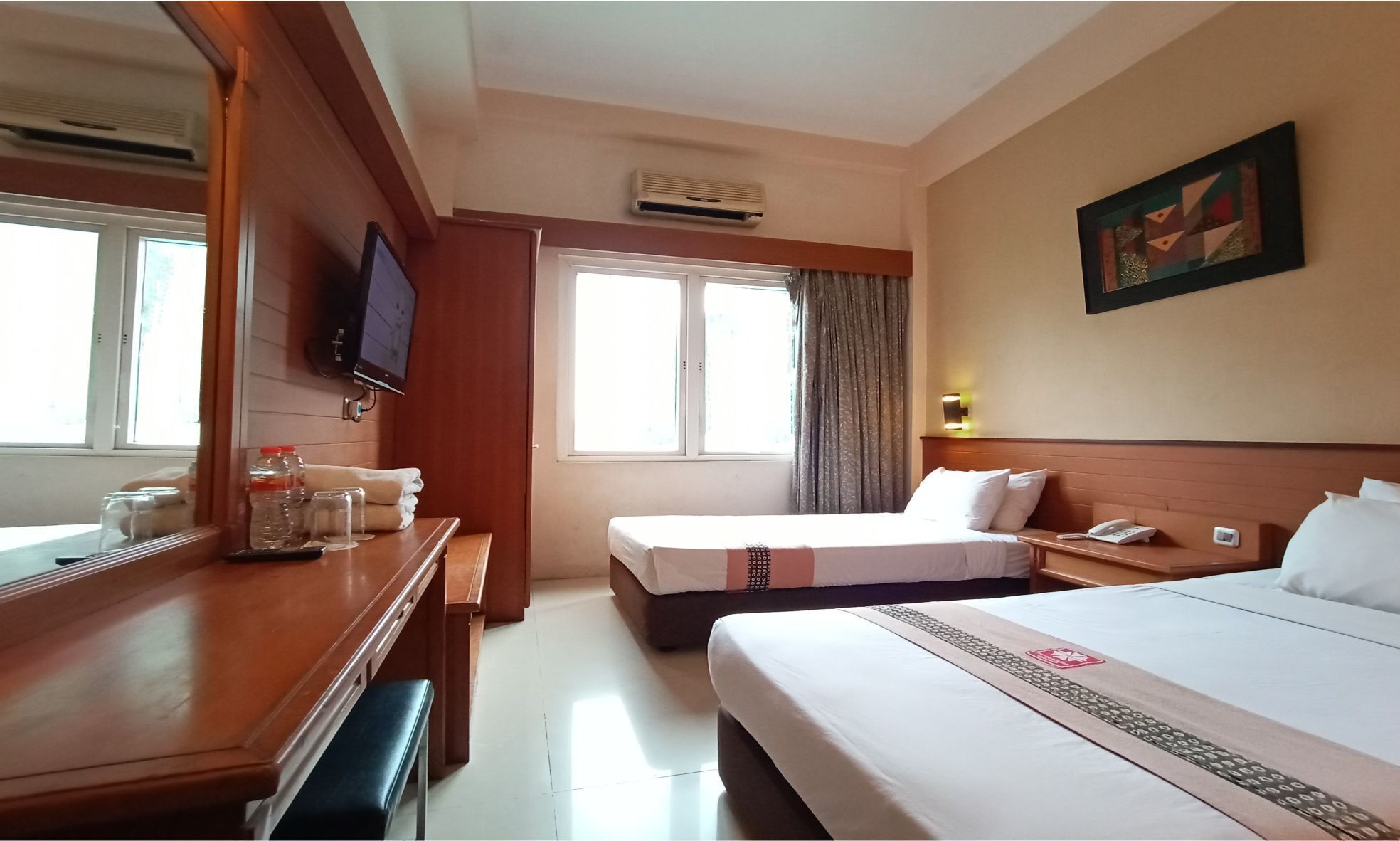A hotel room with two beds, a desk with a television, a window with curtains, and a painting on the wall.