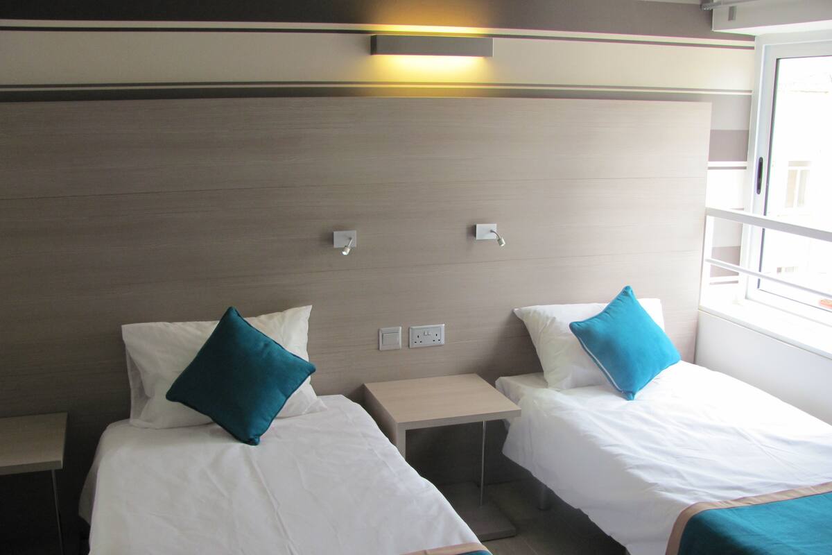 A hotel room with two single beds, each with a teal bedspread and white pillows. There is a small bedside table with a lamp between the beds. The room has a wooden headboard and a window on the right side.