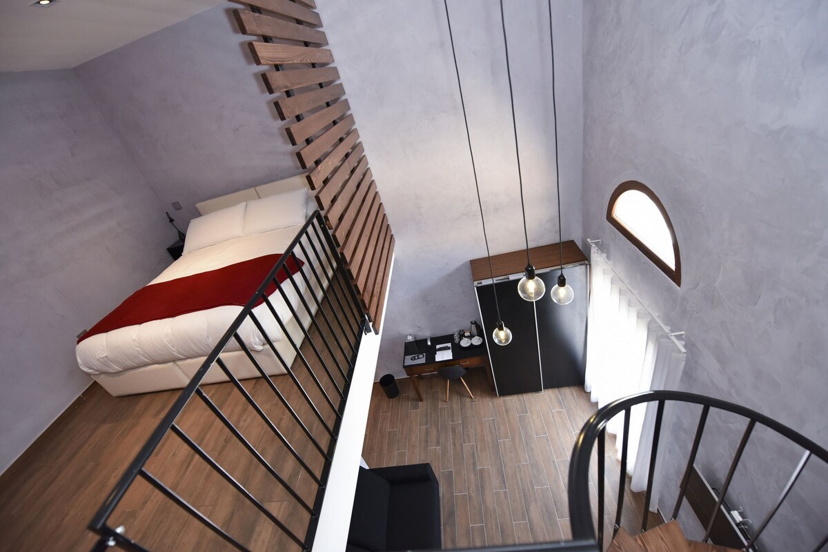 Una habitación moderna con una escalera curva, una cama con una manta roja, un escritorio pequeño con una lámpara y una ventana con vistas al exterior.