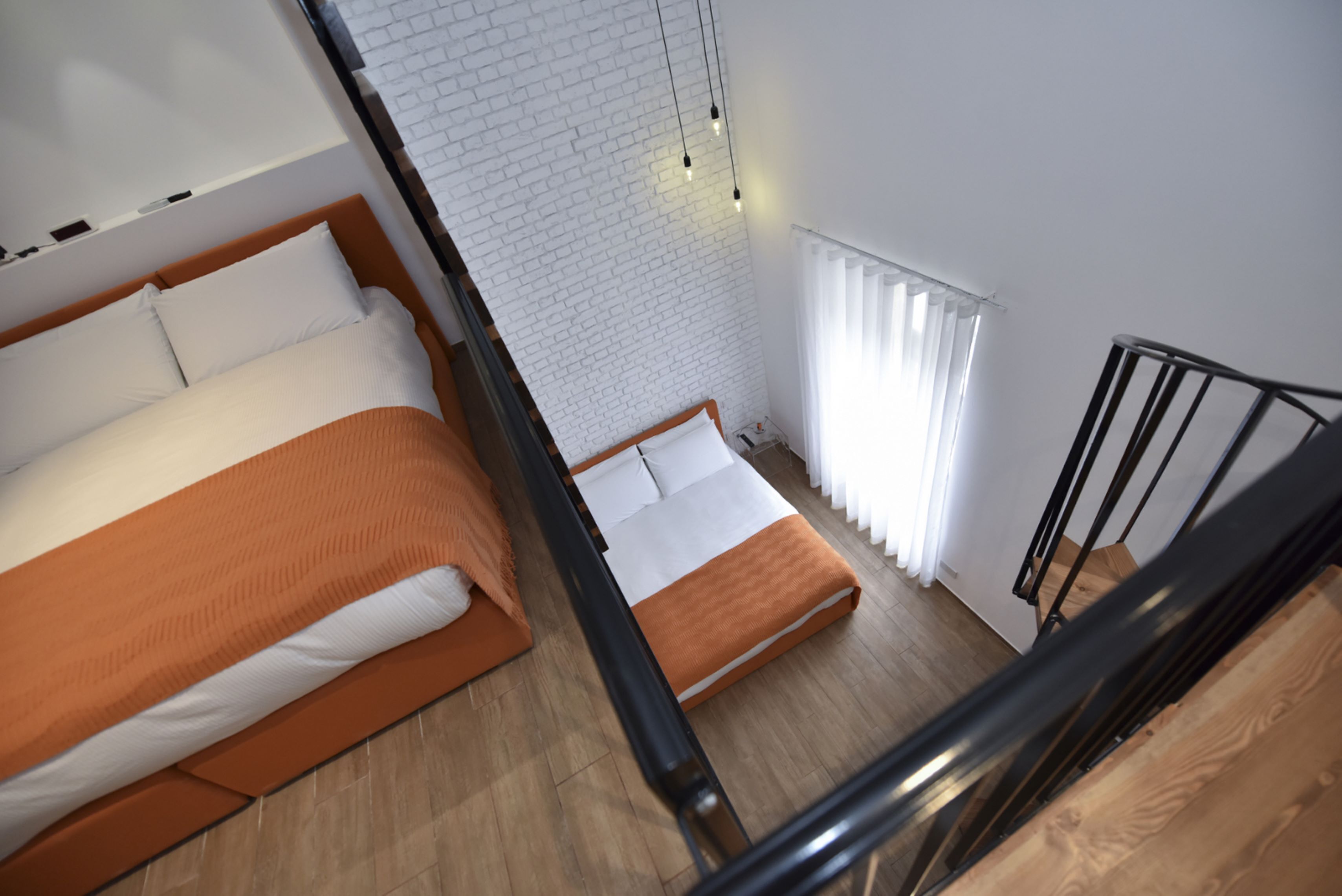 A modern bedroom with a wooden floor, a bed with orange bedding, a white brick wall, and a staircase with a black railing.