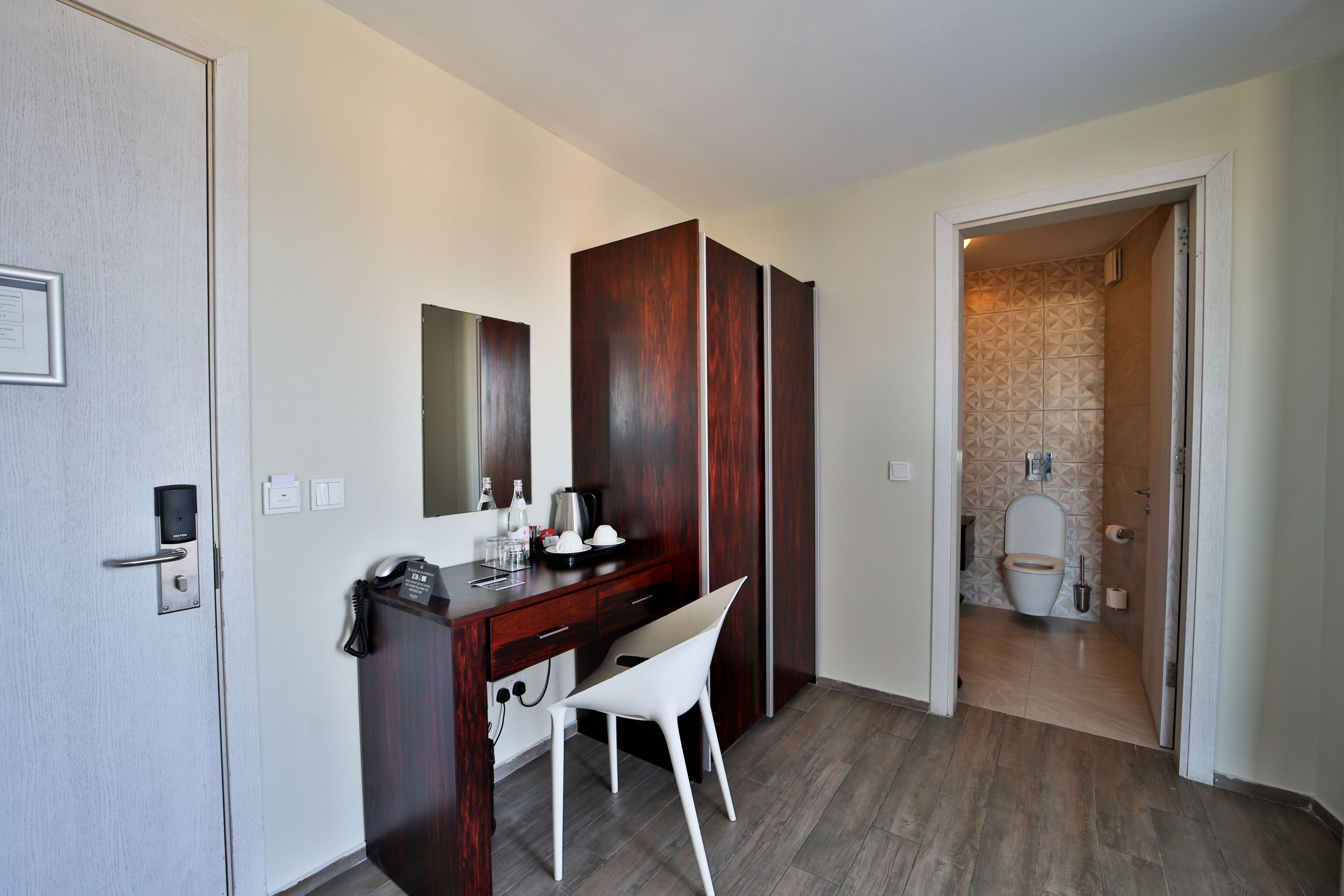 A hotel room with a wooden desk, a white chair, a mirror, and a bathroom visible through an open door.
