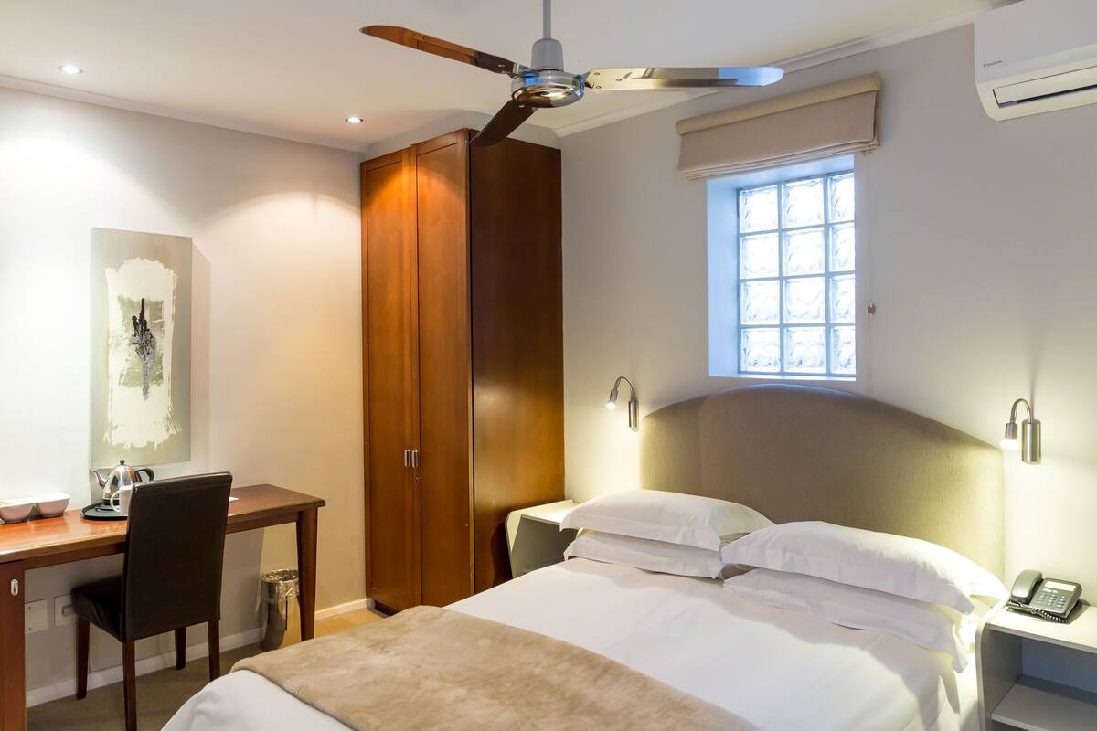Habitación de hotel con cama, escritorio, silla, armario, ventilador de techo, ventana y lámpara de noche.