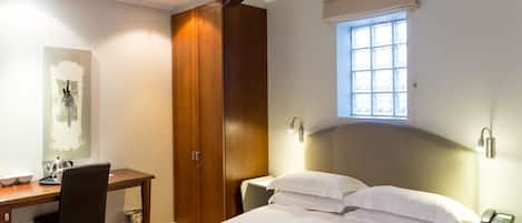 Habitación de hotel con cama, escritorio, silla, armario, ventilador de techo, ventana y lámpara de noche.