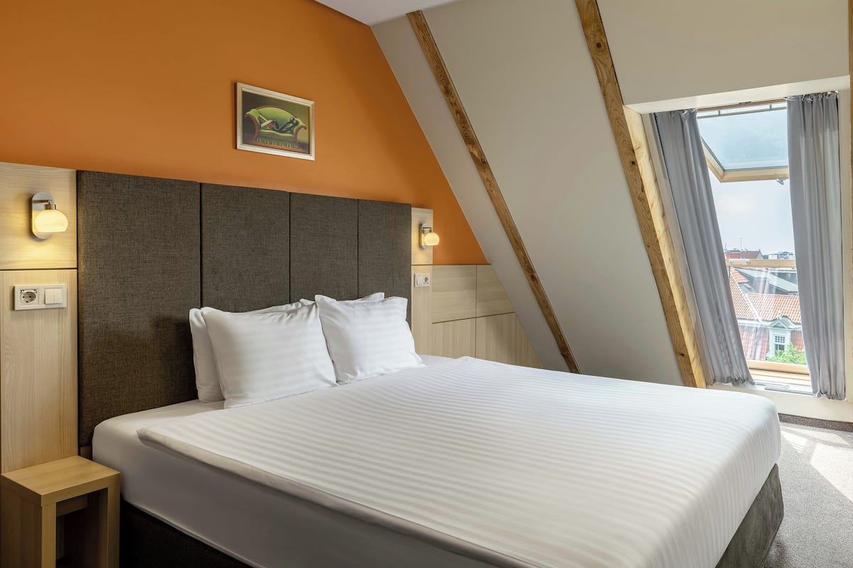 Una habitación de hotel moderna con una cama grande, un cabecero con iluminación integrada, una mesita de noche pequeña y vistas de la ciudad a través de un ventanal amplio.