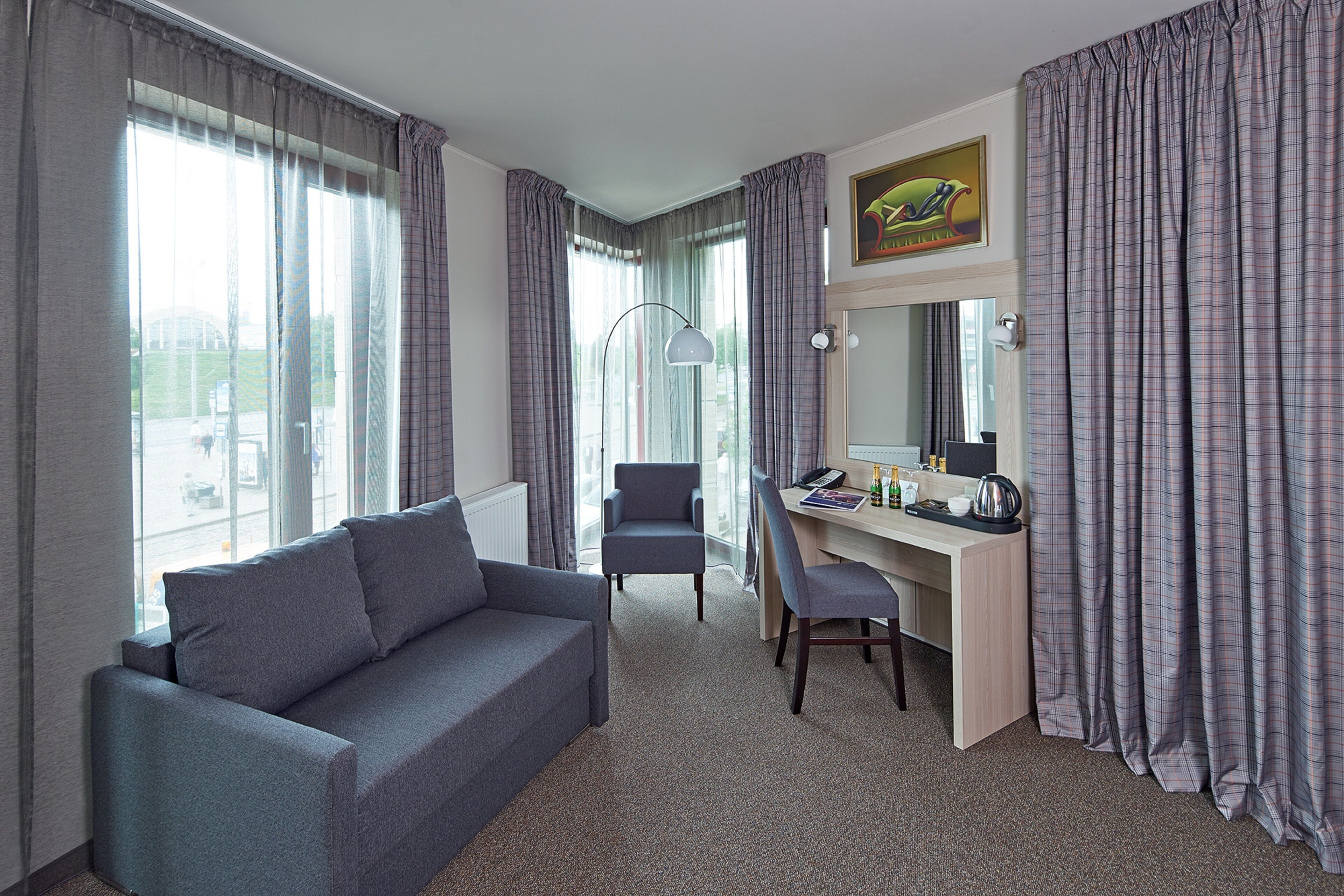 Habitación de hotel con un sofá gris, un escritorio con una silla, una lámpara y un gran ventanal con cortinas.