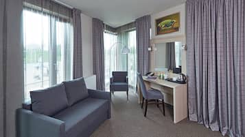 Habitación de hotel con un sofá gris, un escritorio con una silla, una lámpara y un gran ventanal con cortinas.