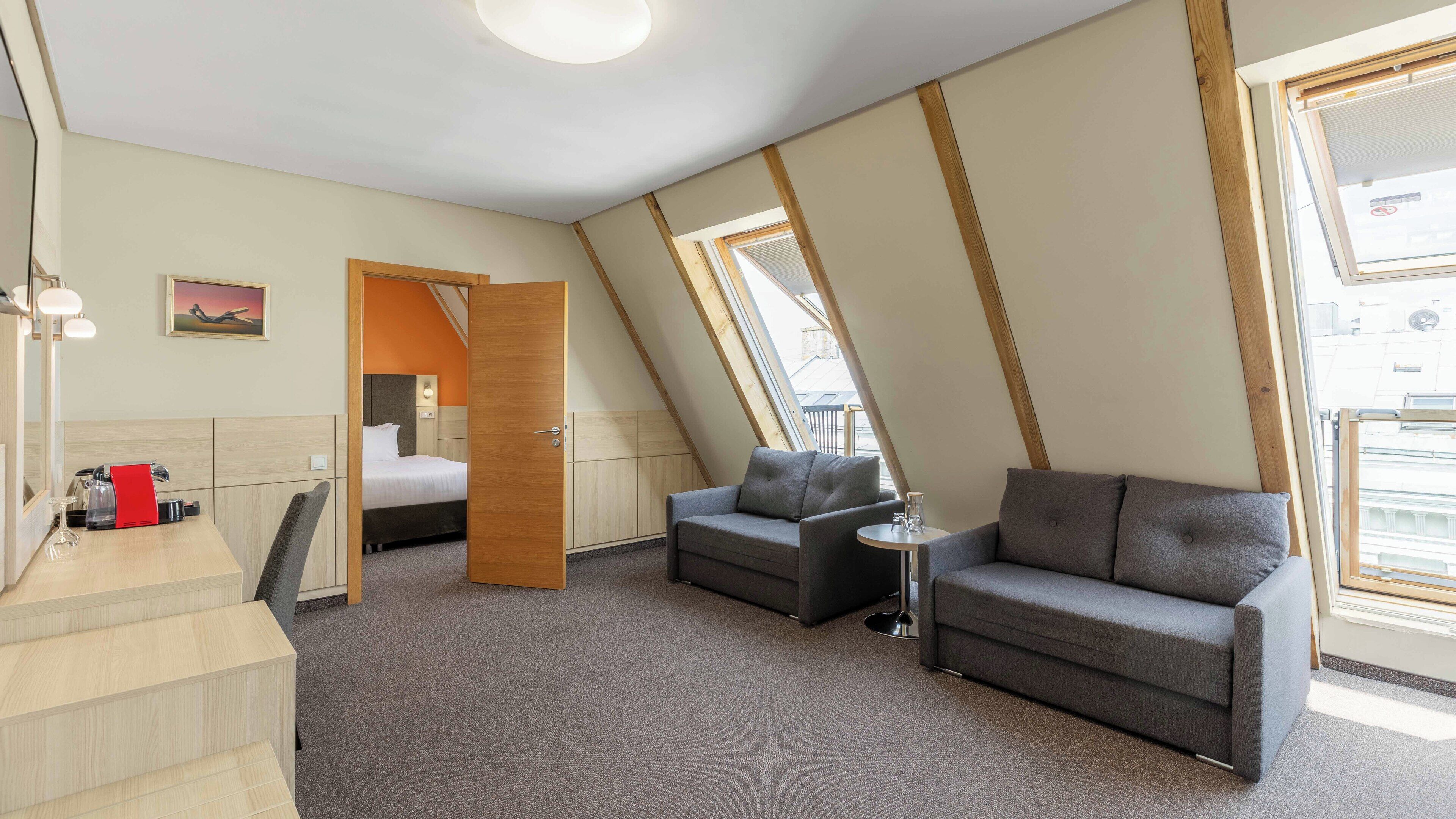 Moderni hotellihuone, jossa on parveltasängy, harmaa sohva, pieni pöytä ja suuri ikkuna, josta avautuu näkymä kaupunkiin.