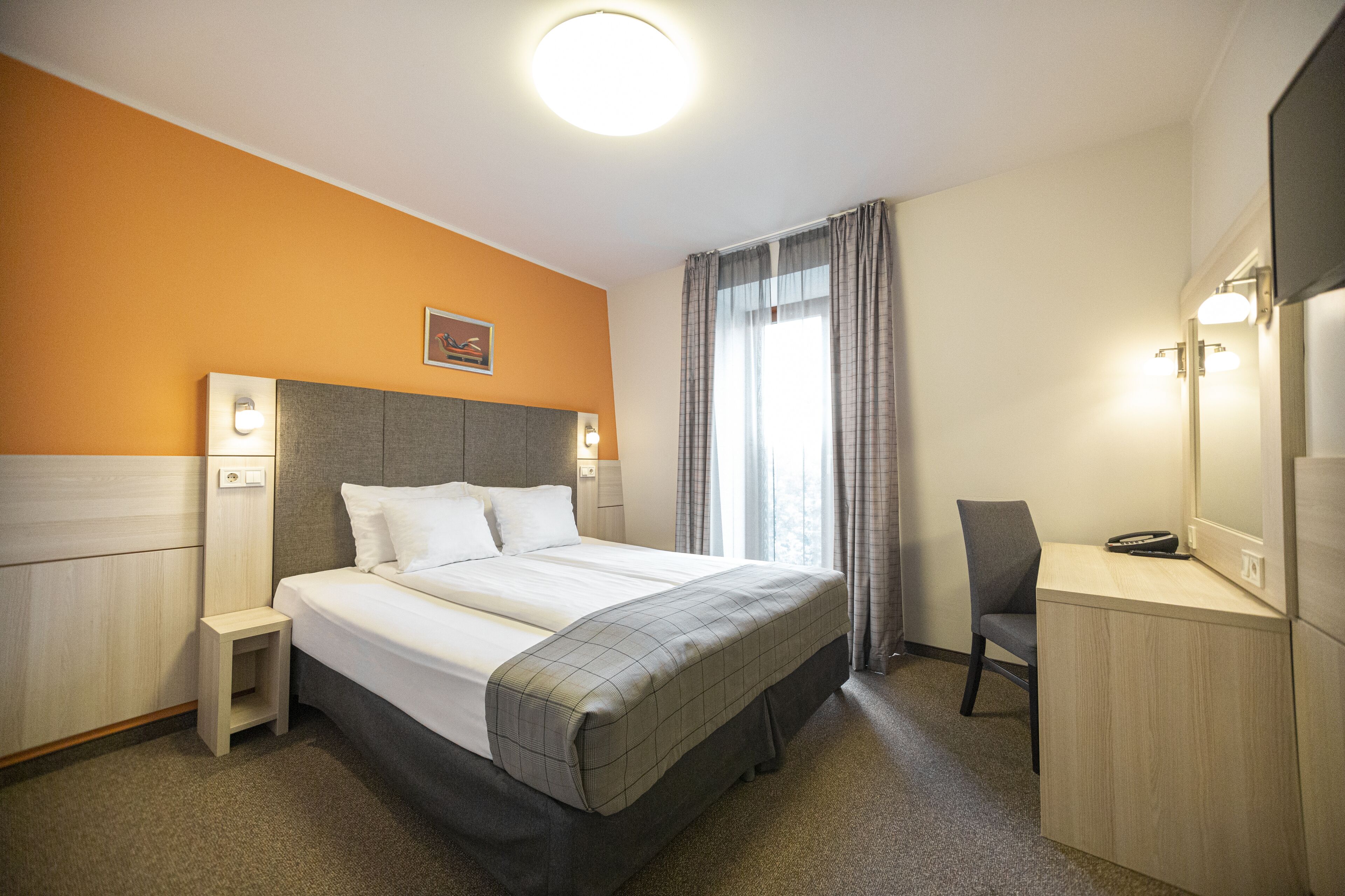 Hotellihuone, jossa on sänky, yöpöytä, tuoli, työpöytä ja peili.