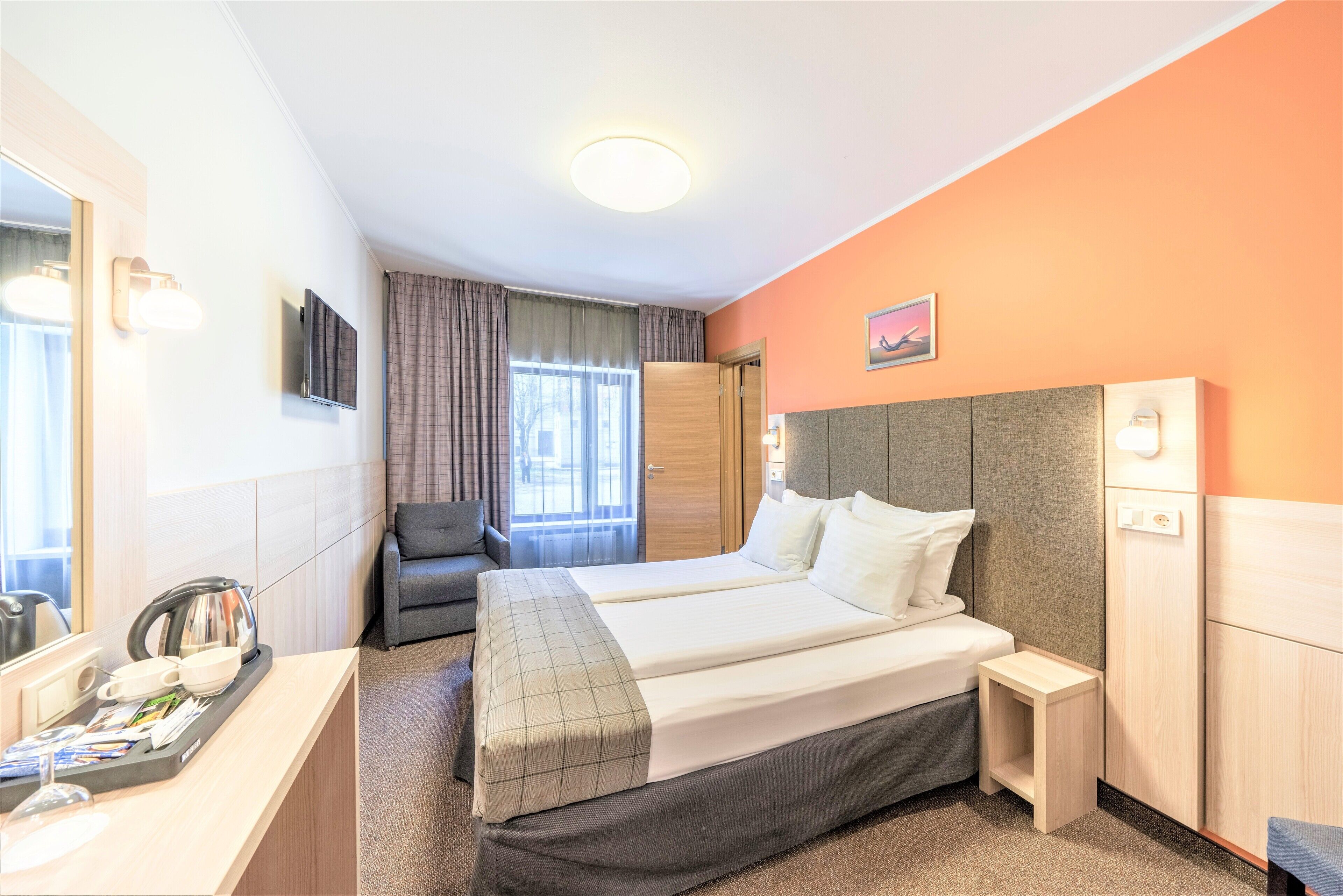 A hotel room with two beds, a desk with a kettle, a chair, and a small bedside table.