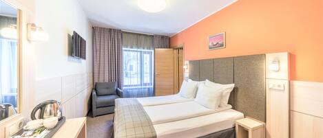 A hotel room with two beds, a desk with a kettle, a chair, and a small bedside table.