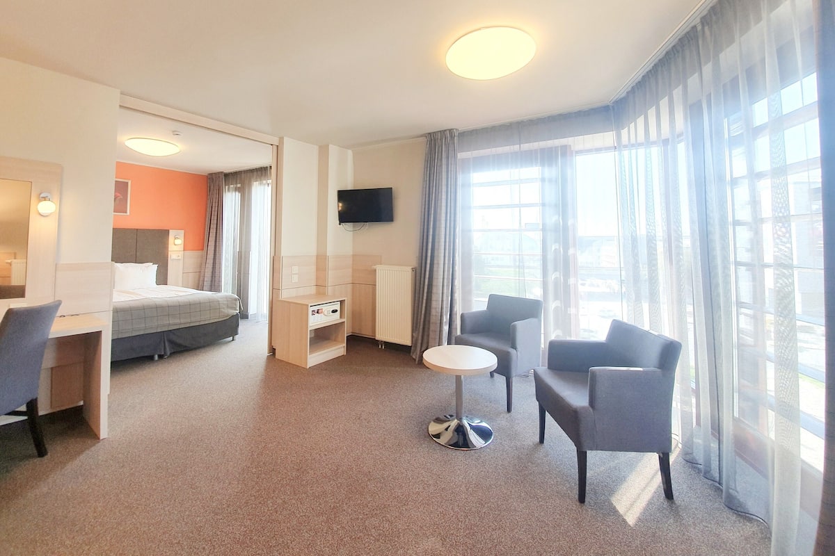 Habitación de hotel con cama, escritorio, silla, televisor y ventanas amplias.