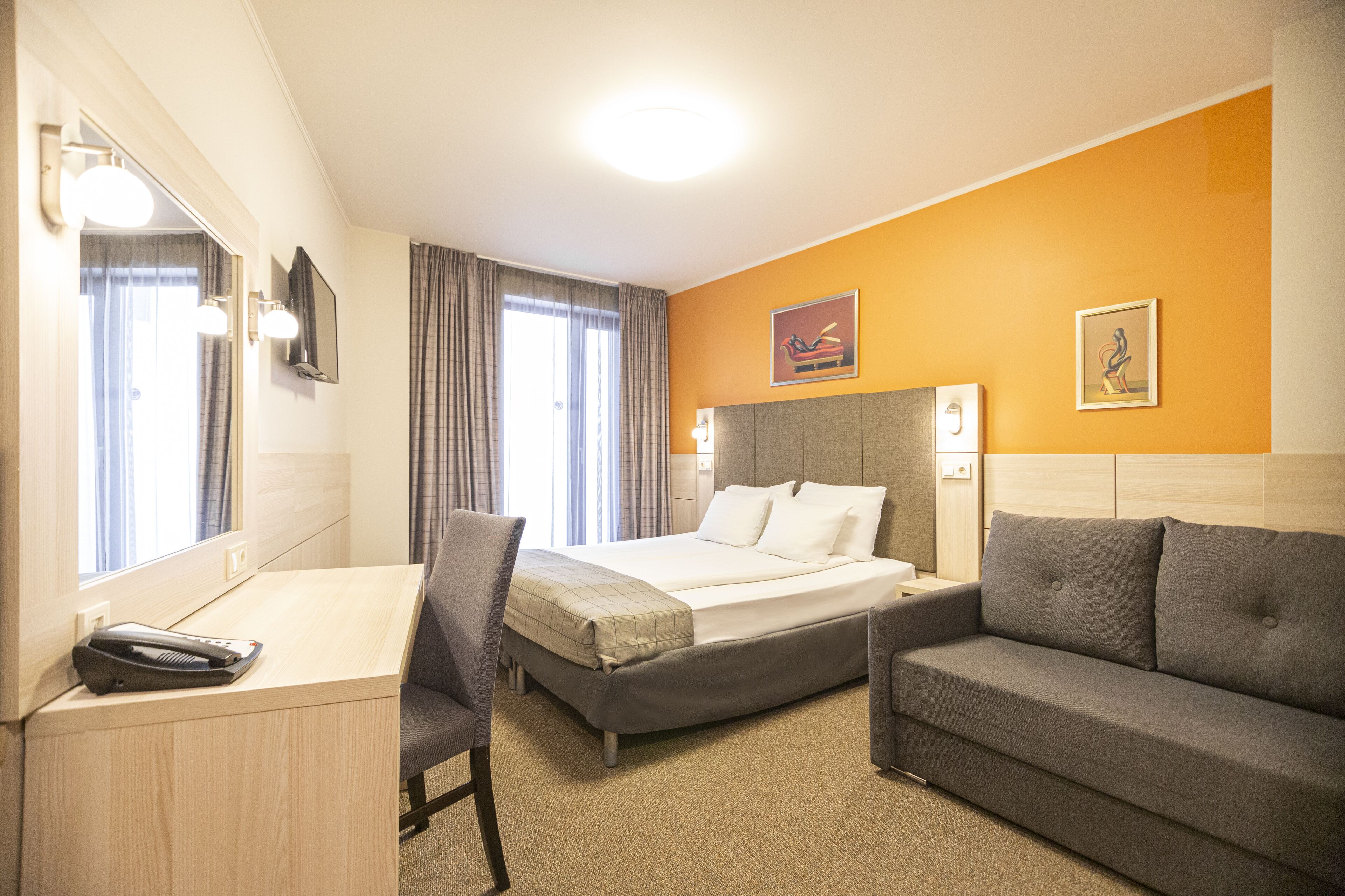 A hotel room with a bed, a desk, a chair, a sofa, a mirror, and two paintings on the wall.