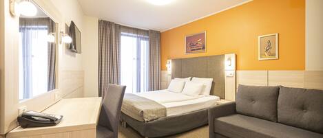 A hotel room with a bed, a desk, a chair, a sofa, a mirror, and two paintings on the wall.
