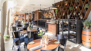 A modern restaurant with round tables, black chairs, and a bar area with shelves of bottles and plants. - Wellton Riga Hotel & SPA (Riga)