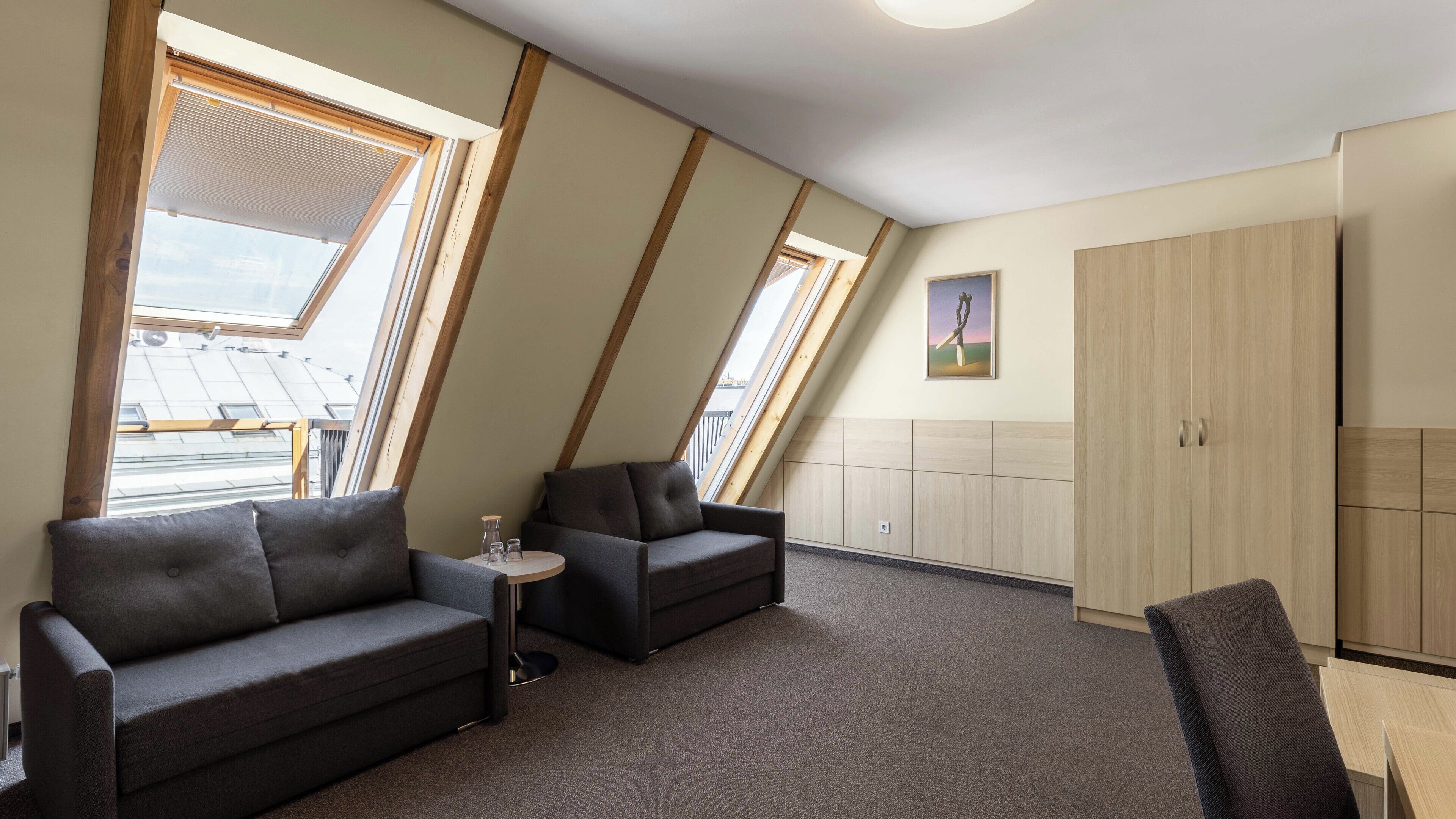 A modern loft bedroom with a sloped ceiling, skylights, a sofa, a chair, and a small table.