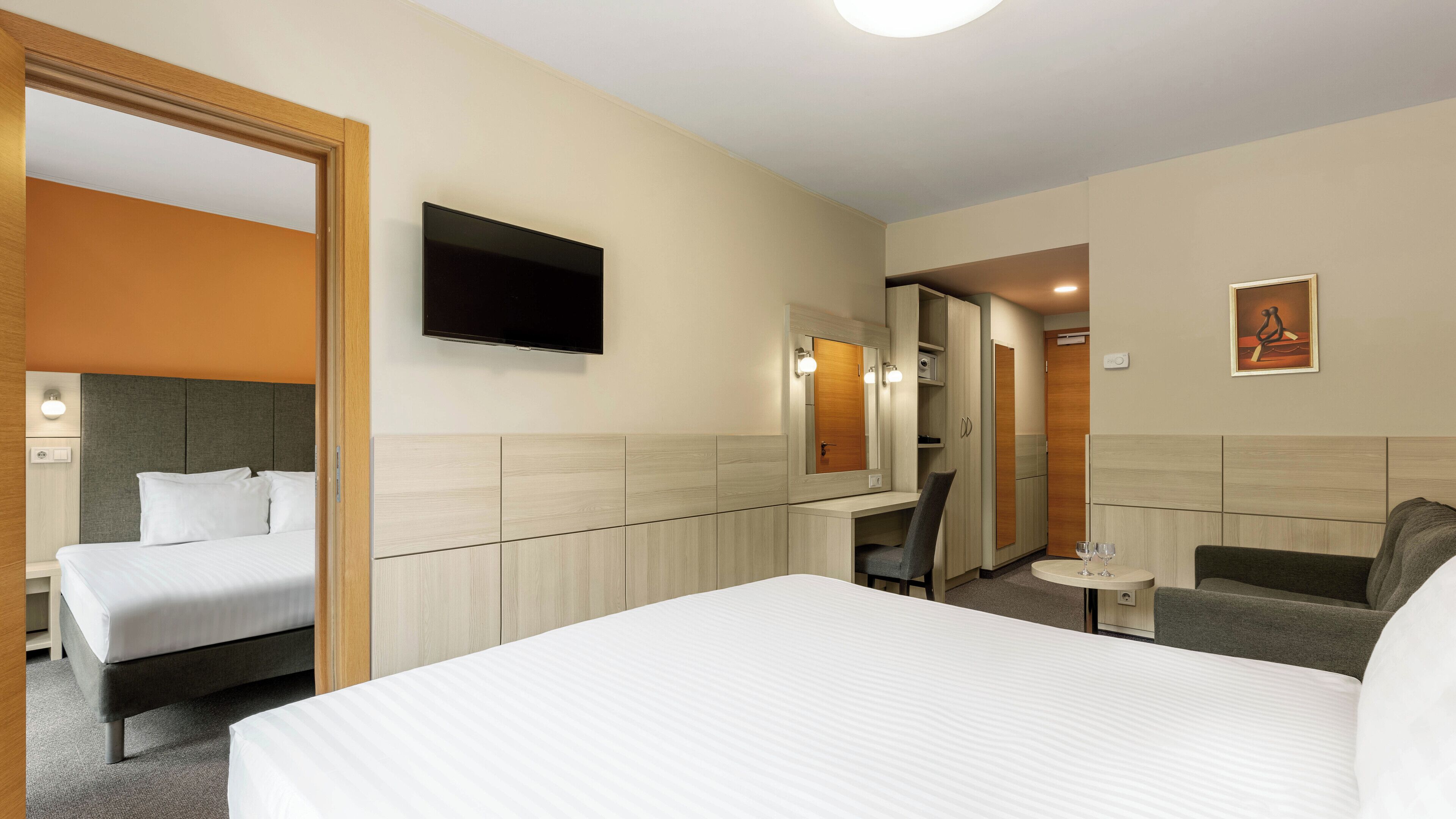 A hotel room with a large bed, a desk, a chair, a TV, and a small table with glasses.
