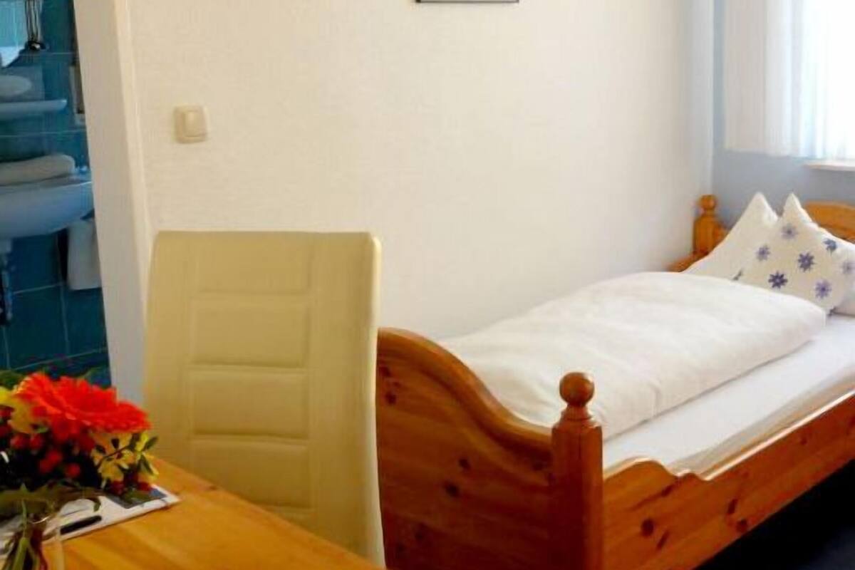 Une petite chambre avec un lit simple, une tête de lit en bois, une table de chevet ornée d’un vase de fleurs et une fen être donnant sur l’extérieur.
