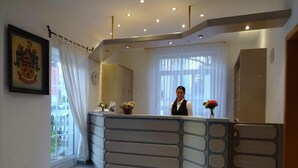 A reception desk with a receptionist, a framed picture on the wall, and a window with curtains. - Hotel Bürgerbräu (Osnabrueck)