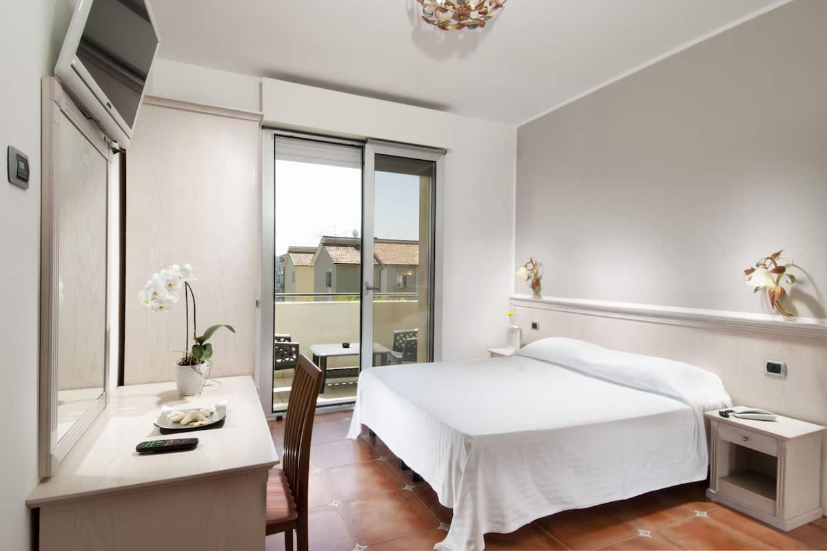 A hotel room with a bed, a desk with a remote control, a chair, a small table, and a balcony with a view of buildings.