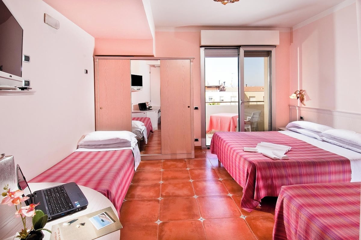 A hotel room with three beds, a laptop on a small table, and a balcony with a table and chairs.