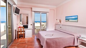 Un dormitorio con cama, escritorio con computadora portátil, silla, televisor y balcón con vista al mar.