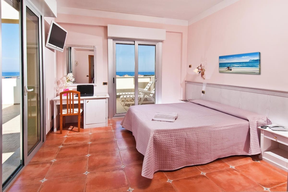 A bedroom with a bed, a desk with a laptop, a chair, a TV, and a balcony with a view of the sea.