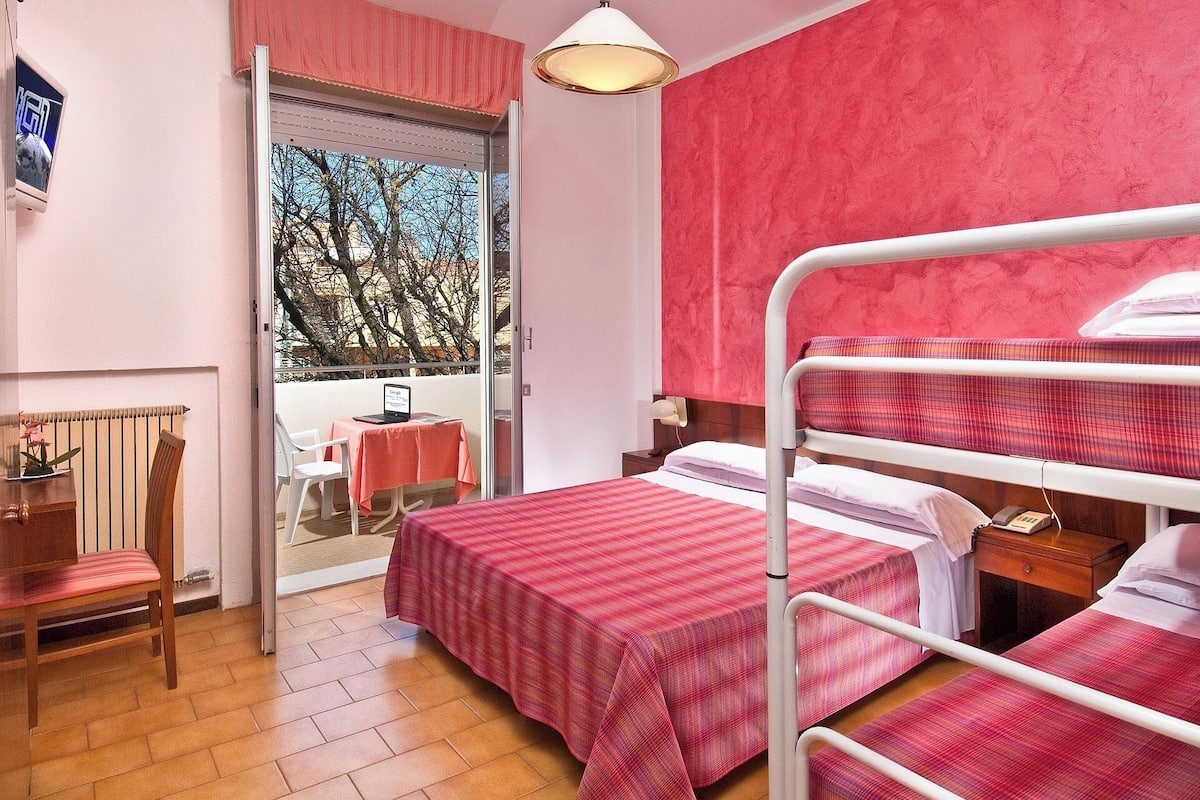 A hotel room with a balcony, a bed with a red checkered bedspread, a wooden chair, and a small table with a laptop.