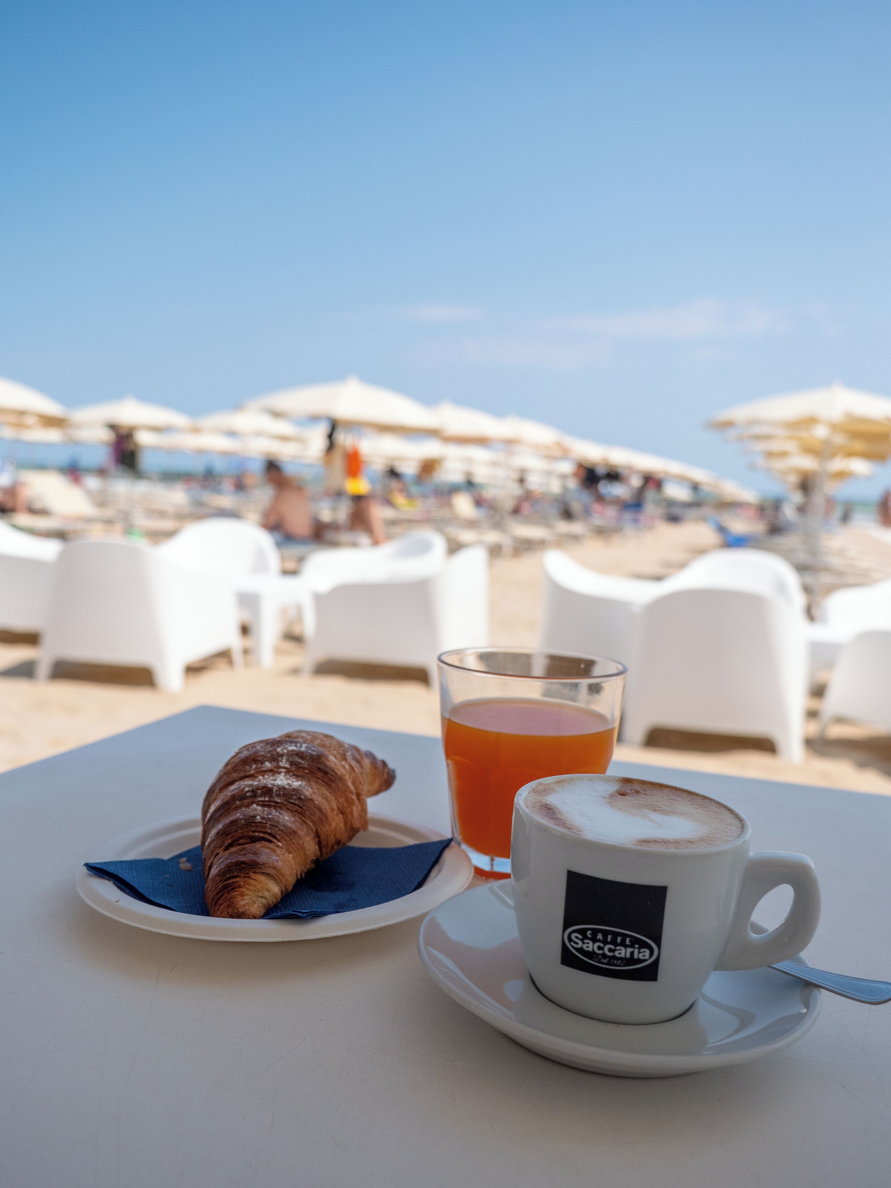 Una mesa con un croissant, un vaso de jugo de naranja y una taza de café con la etiqueta 'SAFFEE SACCARIA'.