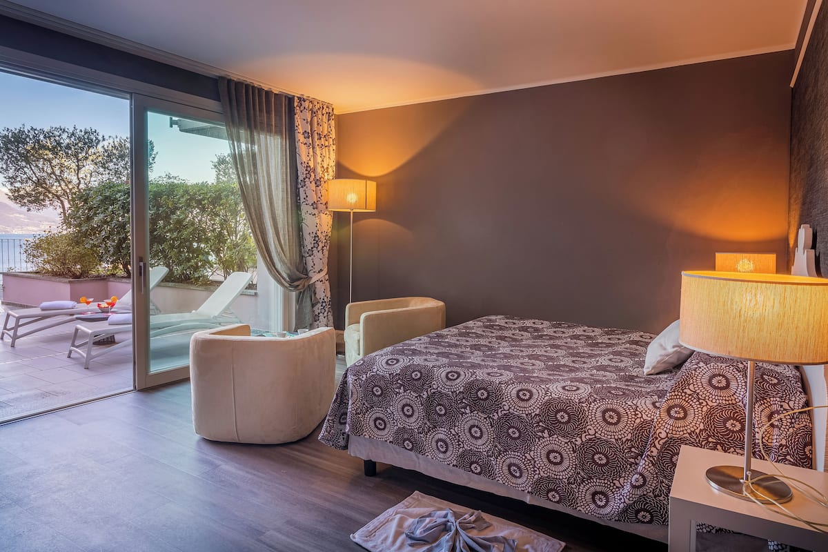 A modern hotel room with a large bed, a sitting area with a chair and a table, a lamp, and a view through a glass door to an outdoor seating area.