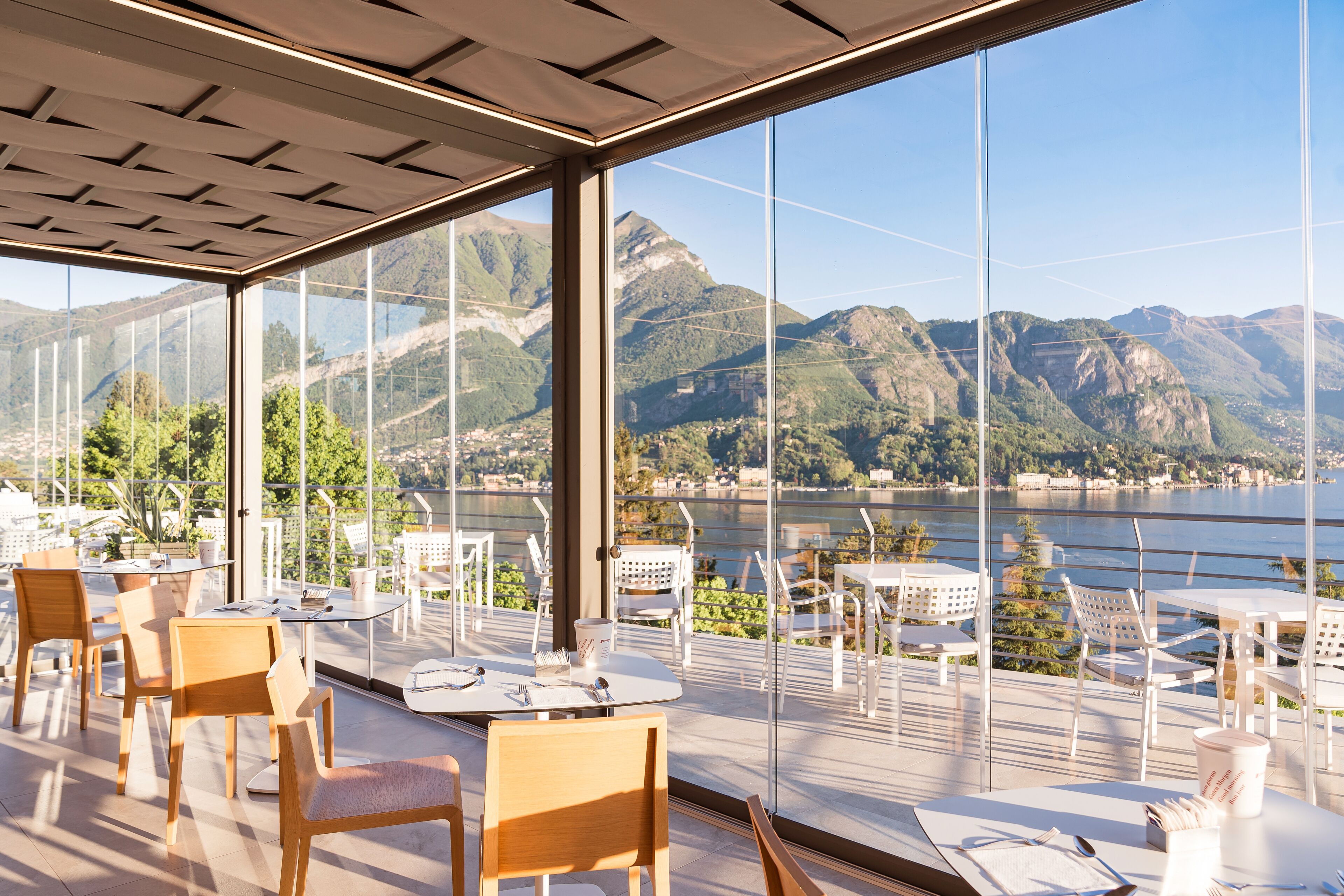 A terrace with tables and chairs, large windows offering a view of mountains and a lake.