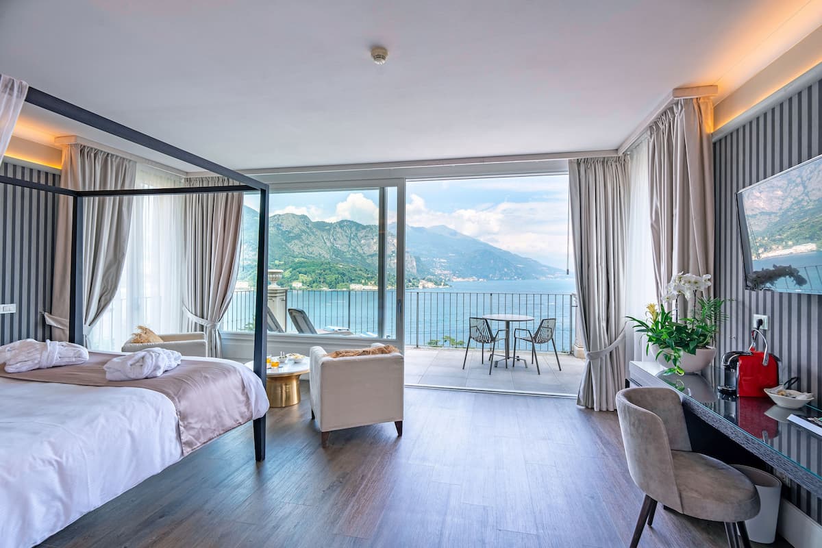 A hotel room with a balcony, a bed, a seating area, and a desk with a view of the sea and mountains.