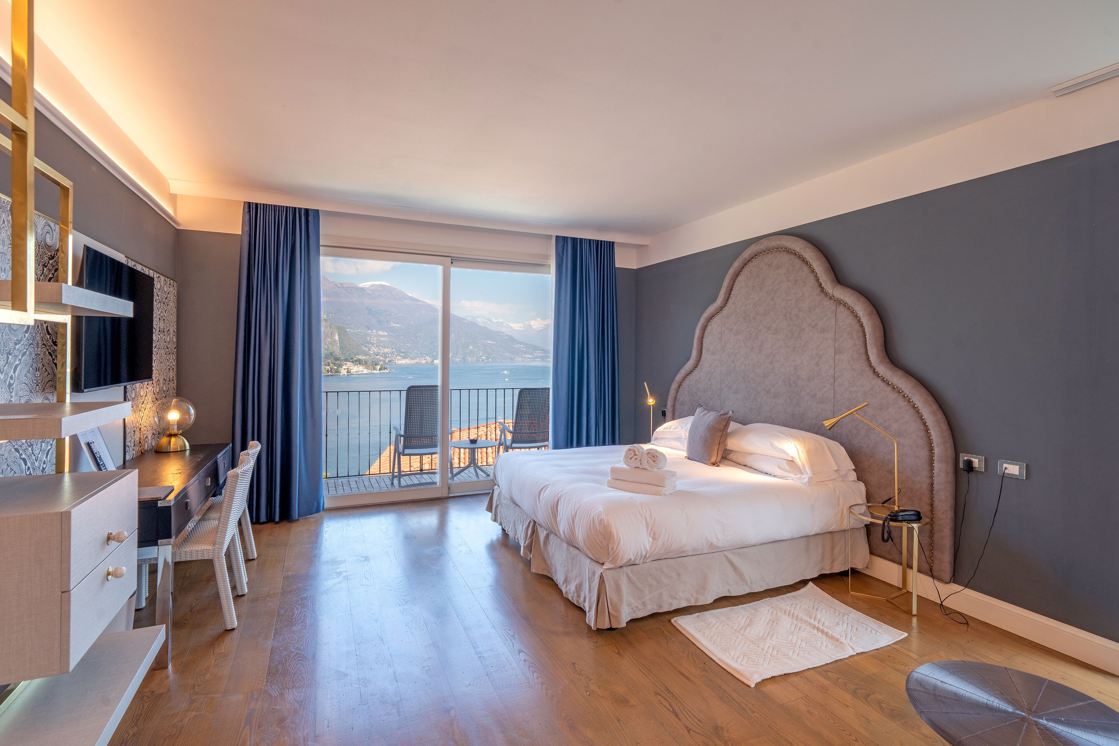 A bedroom with a large bed, a desk with chairs, a balcony with a view of the mountains, and a cityscape.